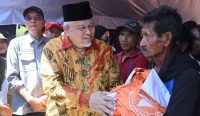 Wakil Ketua Komisi VIII DPR RI, Ansory Siregar, mengenakan batik dan peci hitam, menyerahkan tas bantuan logistik berwarna oranye kepada seorang pria lanjut usia korban gempa bumi di posko pengungsian Kecamatan Reok, Manggarai, Nusa Tenggara Timur.