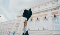 Sebuah tangan mengangkat topi toga kelulusan berwarna hitam dengan rumbai emas, berlatar belakang bangunan megah perguruan tinggi bergaya klasik. Ilustrasi ini merepresentasikan harapan mahasiswa di tengah bergulirnya wacana kuliah gratis PTN yang kini sedang disorot terkait kesiapan anggaran negaranya. (Kredit Foto: RUT MIIT / Unsplash)