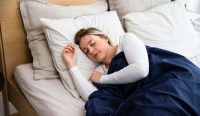 Ilustrasi seorang wanita berambut pirang yang sedang tidur sangat nyenyak di ranjang dengan selimut biru, merepresentasikan tidur kualitas "HD" yang sering dikaitkan dengan tren konsumsi magnesium untuk tidur. (Foto: Slaapwijsheid.nl / Unsplash)