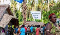Warga mengantre panjang di luar bilik suara sederhana di bawah kibaran bendera daerah saat memberikan hak suara mereka demi Kemerdekaan Bougainville pada referendum 2019, sebuah langkah bersejarah untuk lepas dari Papua Nugini. (Kredit Foto: Ness Kerton/AFP/Getty Images)