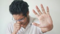 Pria berkacamata sedang batuk sambil mengulurkan tangan memberi isyarat berhenti, sebagai ilustrasi menolak mitos kehujanan bikin flu. (Kredit Foto: Towfiqu Barbhuiya/Unsplash)