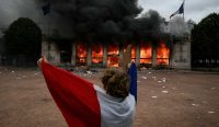 Seorang pengunjuk rasa berbalut bendera nasional berdiri menyaksikan kantor pariwisata Lyon yang terbakar hebat di tengah Kerusuhan Prancis. (Foto: Jeff Pachoud/AFP/Getty Images)