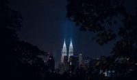 Pemandangan malam Menara Kembar Petronas di Kuala Lumpur yang dibingkai oleh siluet pepohonan gelap. Gedung-gedung tinggi di kawasan urban Malaysia sangat diwajibkan menggunakan penangkal petir standar untuk menghindari ancaman mematikan dari kebakaran akibat petir selama cuaca ekstrem. (Kredit Foto: Muhammad Faiz Zulkeflee/Unsplash)