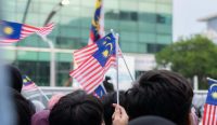 Ilustrasi kerumunan warga Malaysia mengibarkan bendera Jalur Gemilang, merepresentasikan sorotan publik terhadap Kasus Bullying Sekolah Malaysia yang mencapai 4.000 insiden dalam enam bulan. Kredit Foto: Putra Mahirudin / Unsplash.