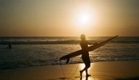 Siluet seorang peselancar membawa papan selancar di Pantai Canggu, Kabupaten Badung, dengan latar matahari senja yang cerah. Kondisi cuaca pesisir yang hangat ini sangat mendukung aktivitas liburan sesuai prakiraan cuaca bali akhir pekan dari BMKG. (Foto: Ivan Torres/Unsplash).